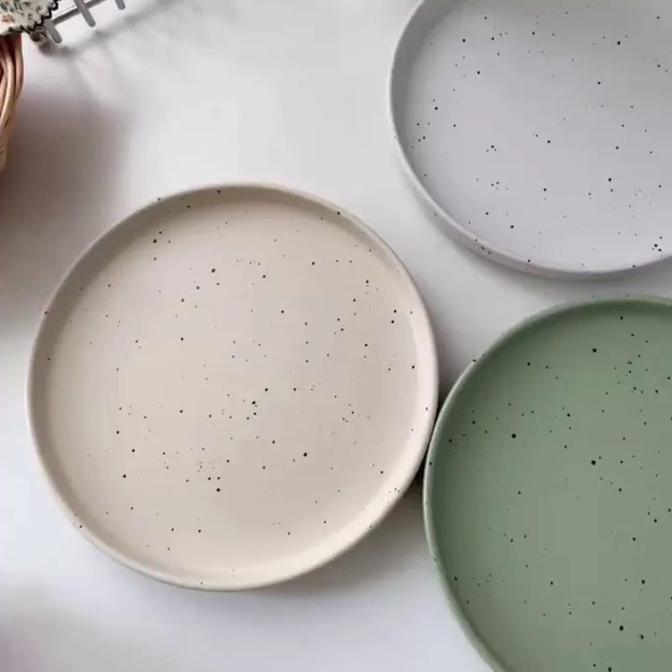 Three minimalist speckled ceramic plates in cream, light grey, and sage green
