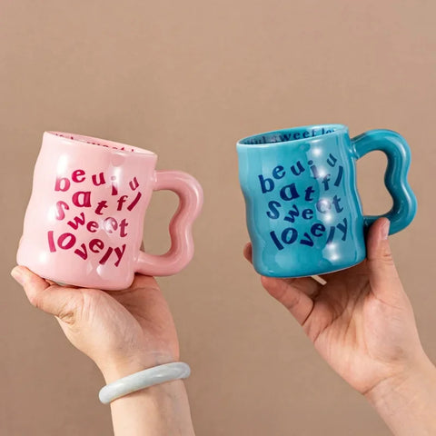 Pink and blue aesthetic wavy ceramic mugs with quirky "beautiful, sweet, lovely" text, held by hands against a neutral background.